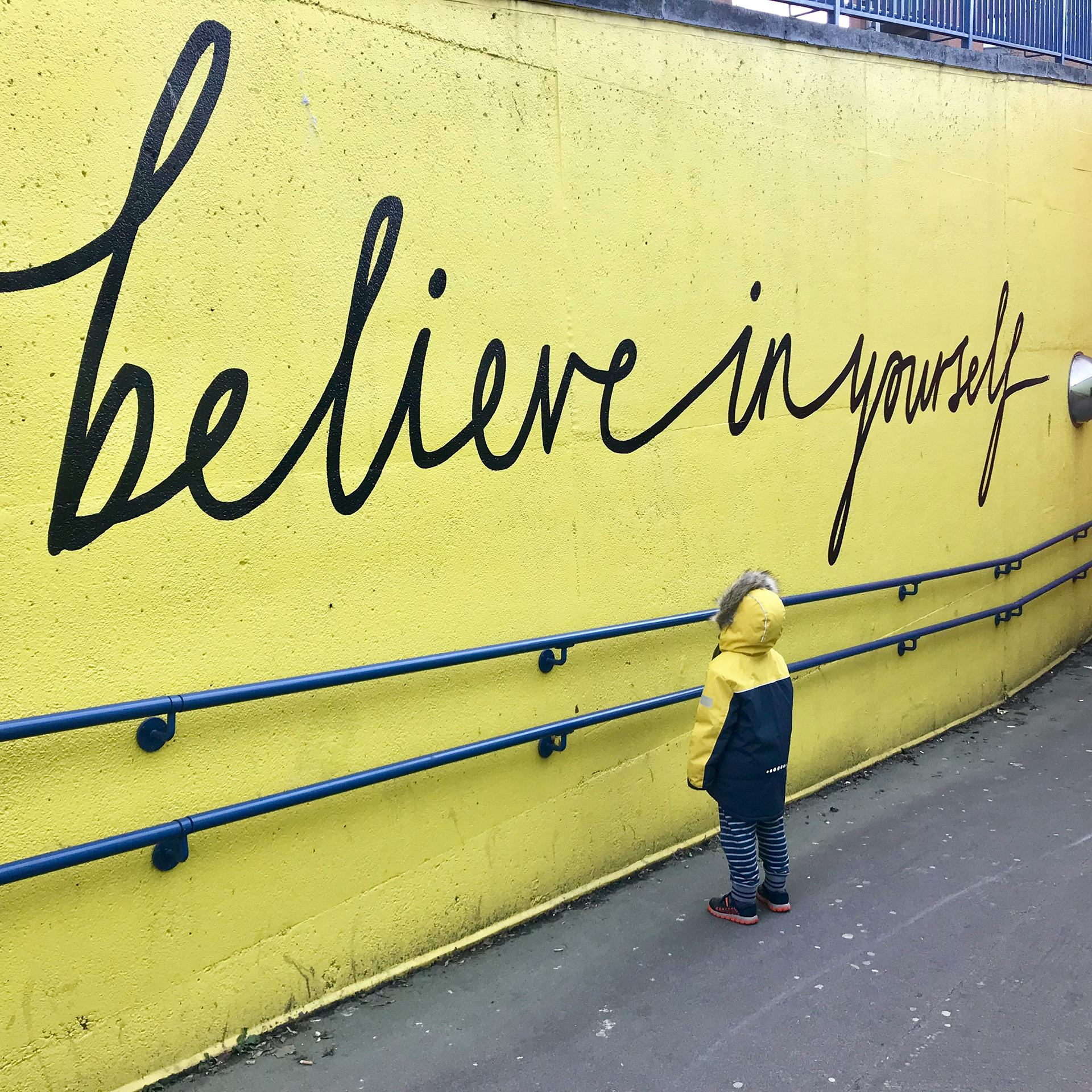 enfant devant un mur jaune avec inscrit dessus "believe in yourself"